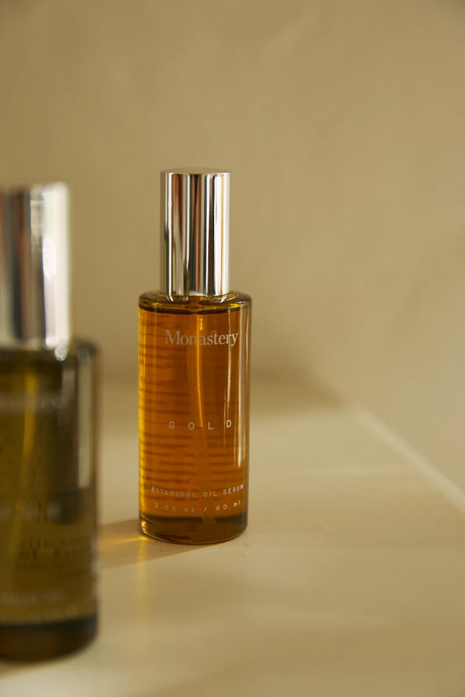 Monastery Gold botanical oil serum bottle with silver cap against neutral background. Amber oil visible, another bottle blurred in foreground.