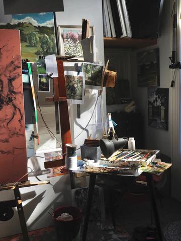 Sunlit art studio with easel holding unfinished paintings. Table covered in paint supplies. Photos and small artworks pinned to board nearby.