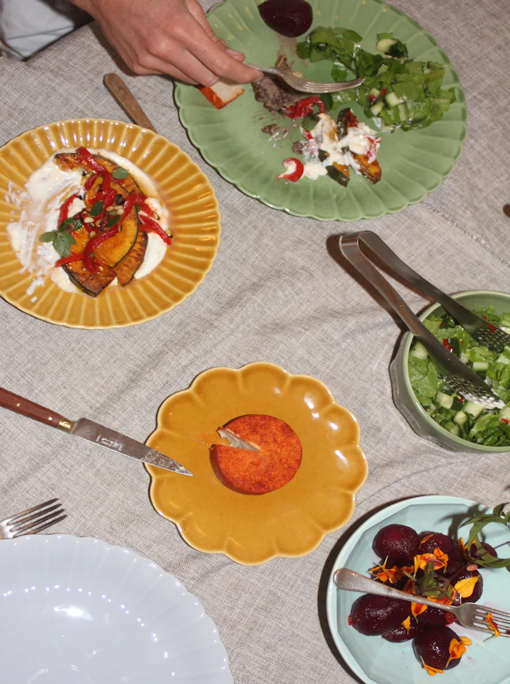 Vibrant table setting with colourful plates of roasted veg, salad, and beets. Hand with fork reaching for food. Neutral tablecloth with flowers, utensils visible.