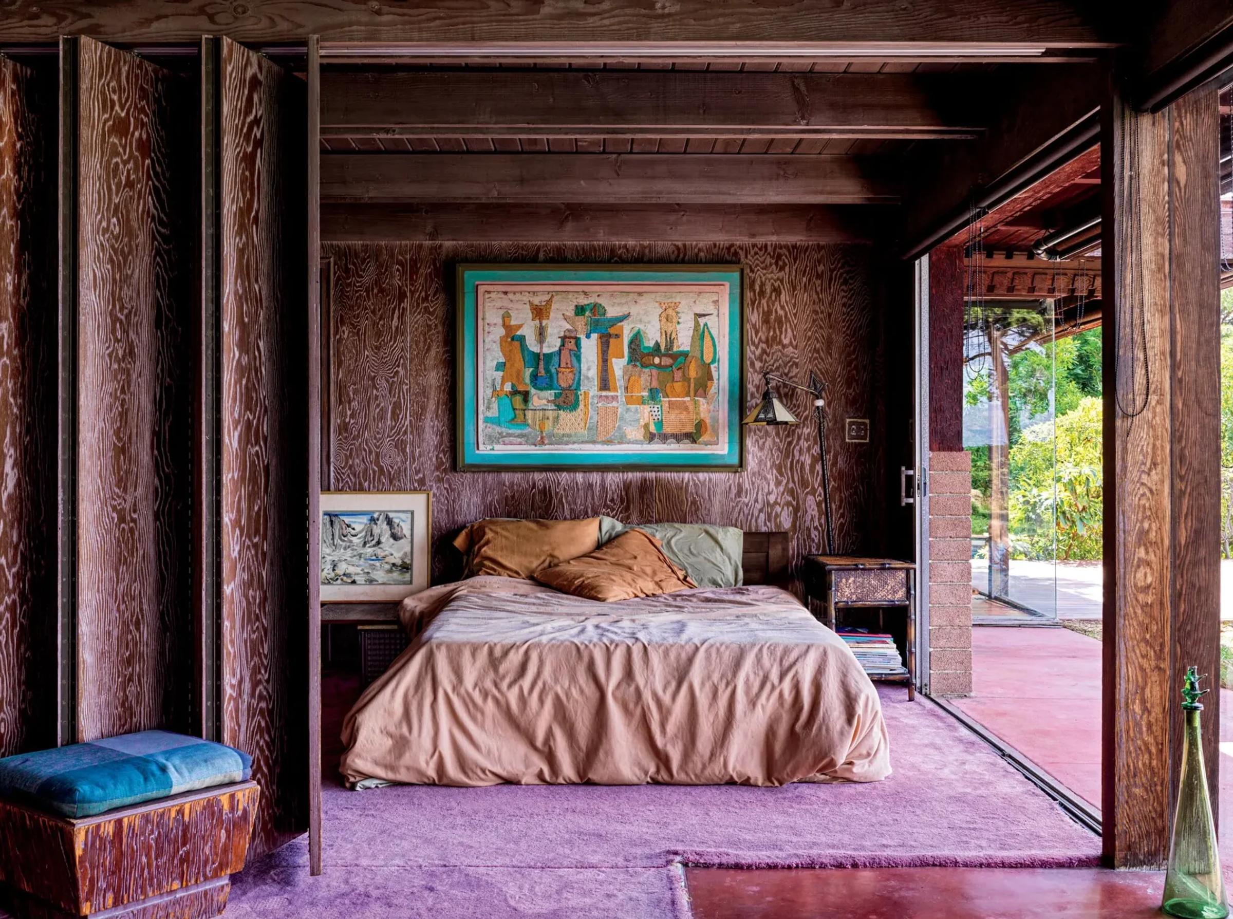 A rustic bedroom with wooden walls with a bed with beige and brown bedding. A colourful artwork hangs above it, with a ceiling height glass slider on the right, showing greenery outside.