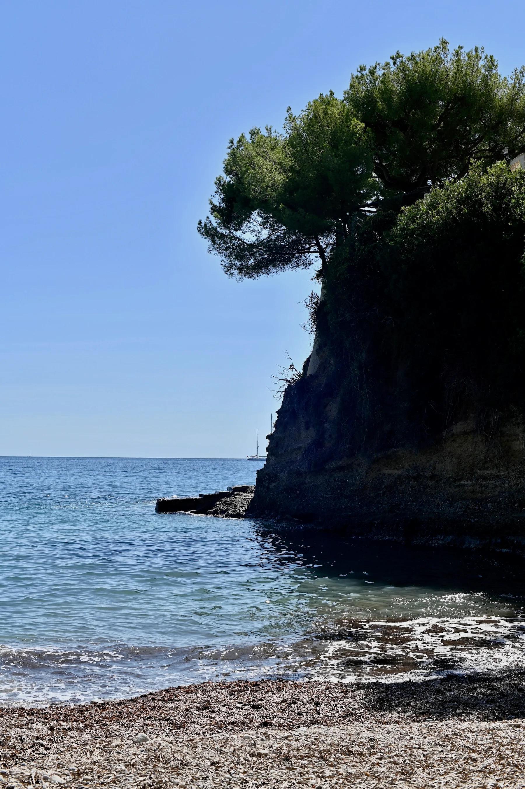 Coastal view with rocky shoreline and clear blue ocean. Tall cliff with green trees extends into water. Clear, bright sky.