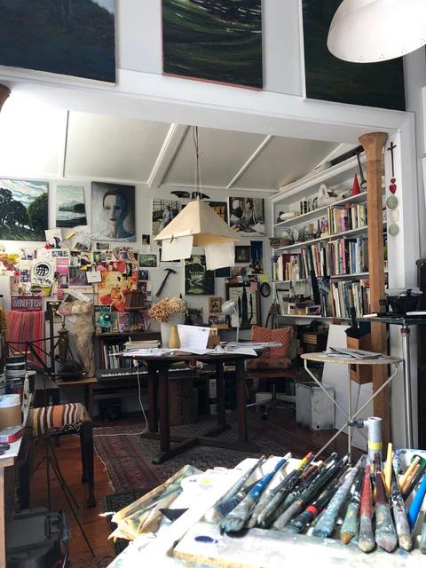 Bright studio filled with Max Thomson's paintings. Bookshelf and table with art supplies. Large windows illuminate room.