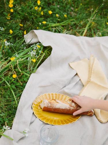 A hand reaches for a baguette on a yellow plate on a white picnic blanket in a greassy field with buttercups. There is also a napkin and a clear plastic cup.