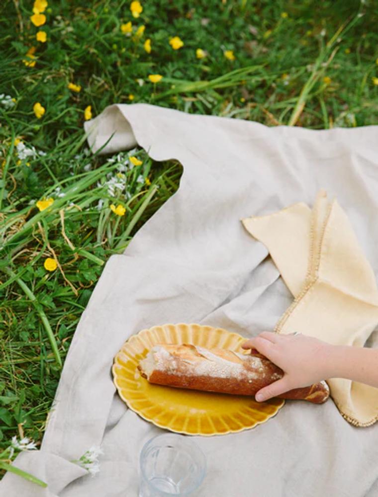 A hand reaches for a baguette on a yellow plate on a white picnic blanket in a greassy field with buttercups. There is also a napkin and a clear plastic cup.