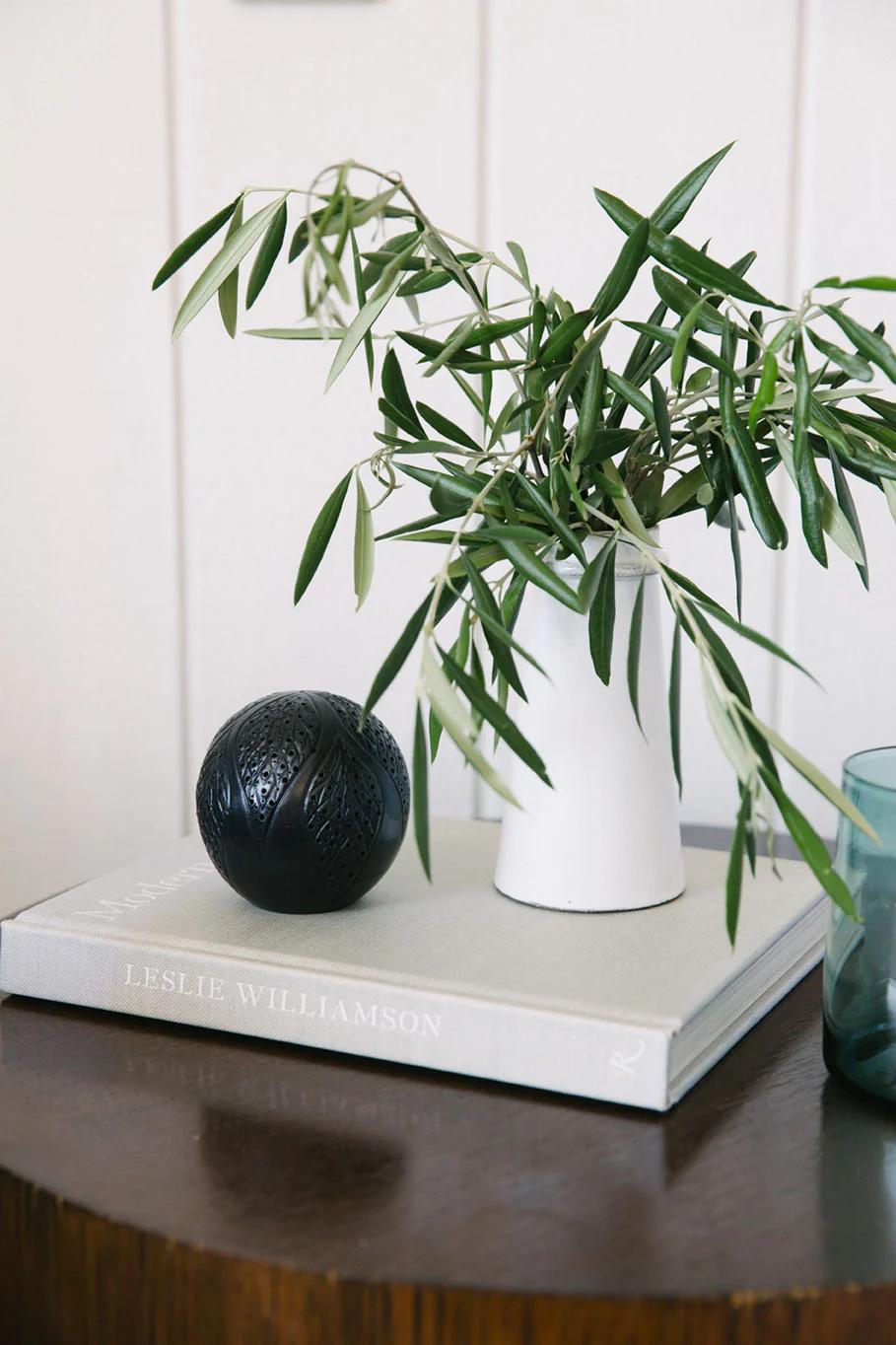 White vase with green branches on "Leslie Williamson" book. Black sphere and teal glass nearby. Dark wood table, light wall background.