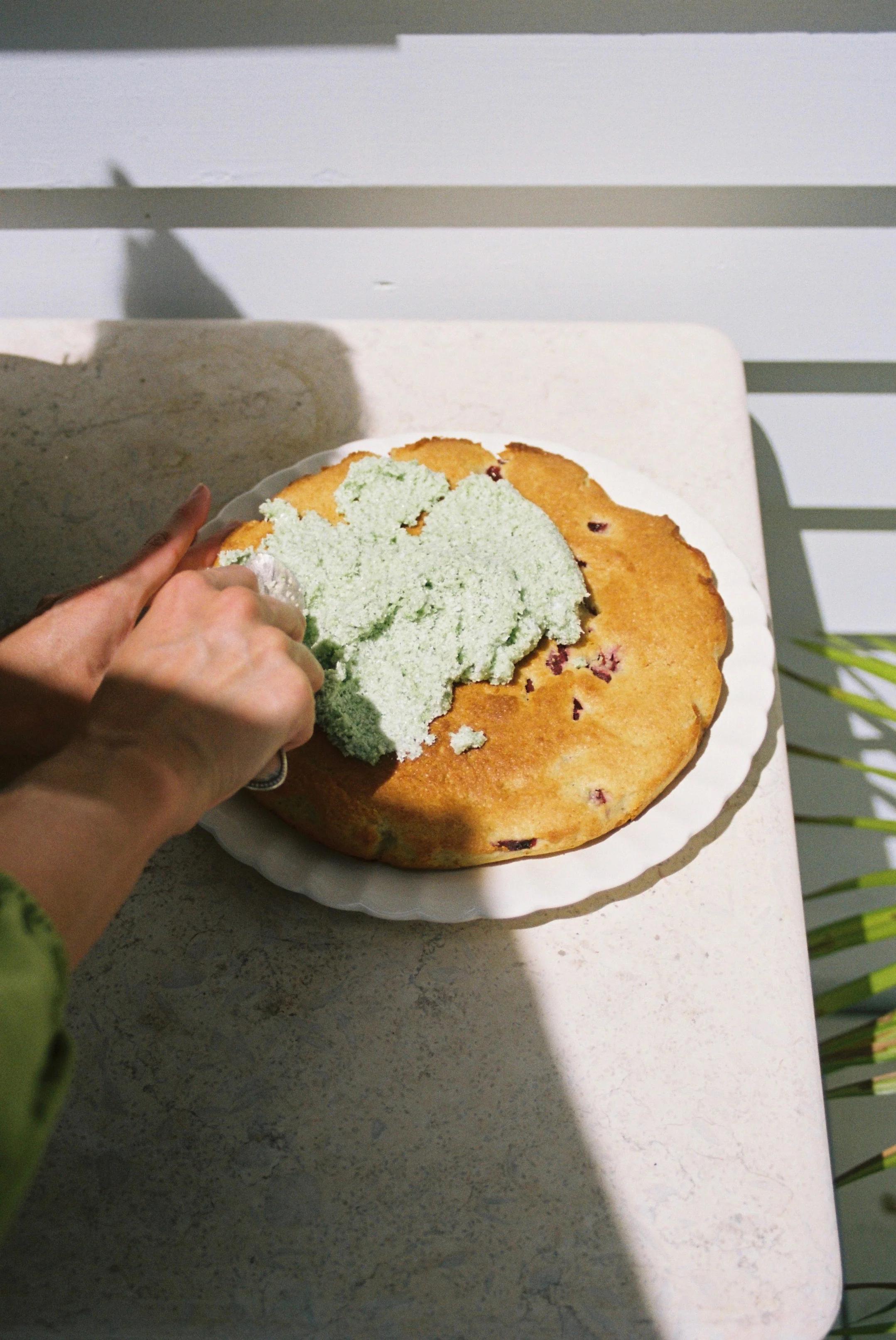 Person spreading green frosting on fruit-topped cake. White plate on light tabletop, shadows from foliage visible.