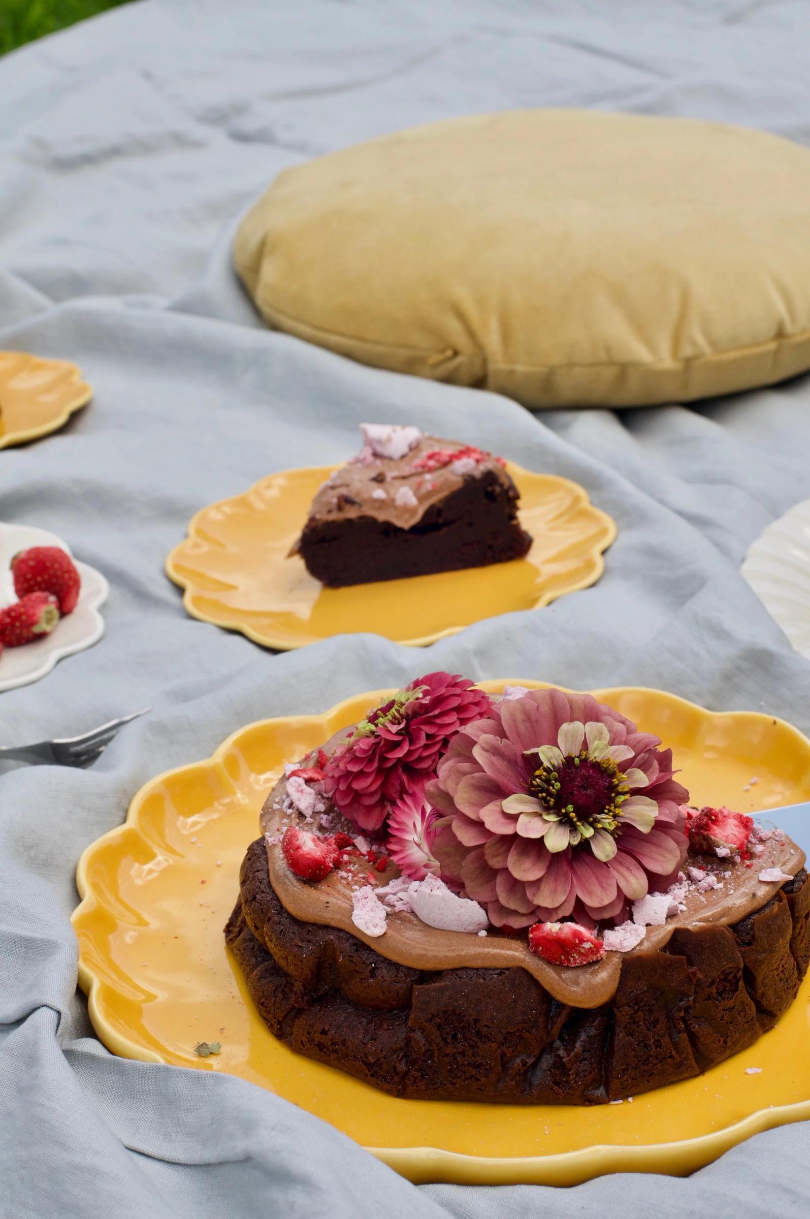 A chocolate cake from The Caker, topped with flowers and strawberries, sits on a yellow plate. A slice of cake and raspberries are placed on a smaller matching plate.