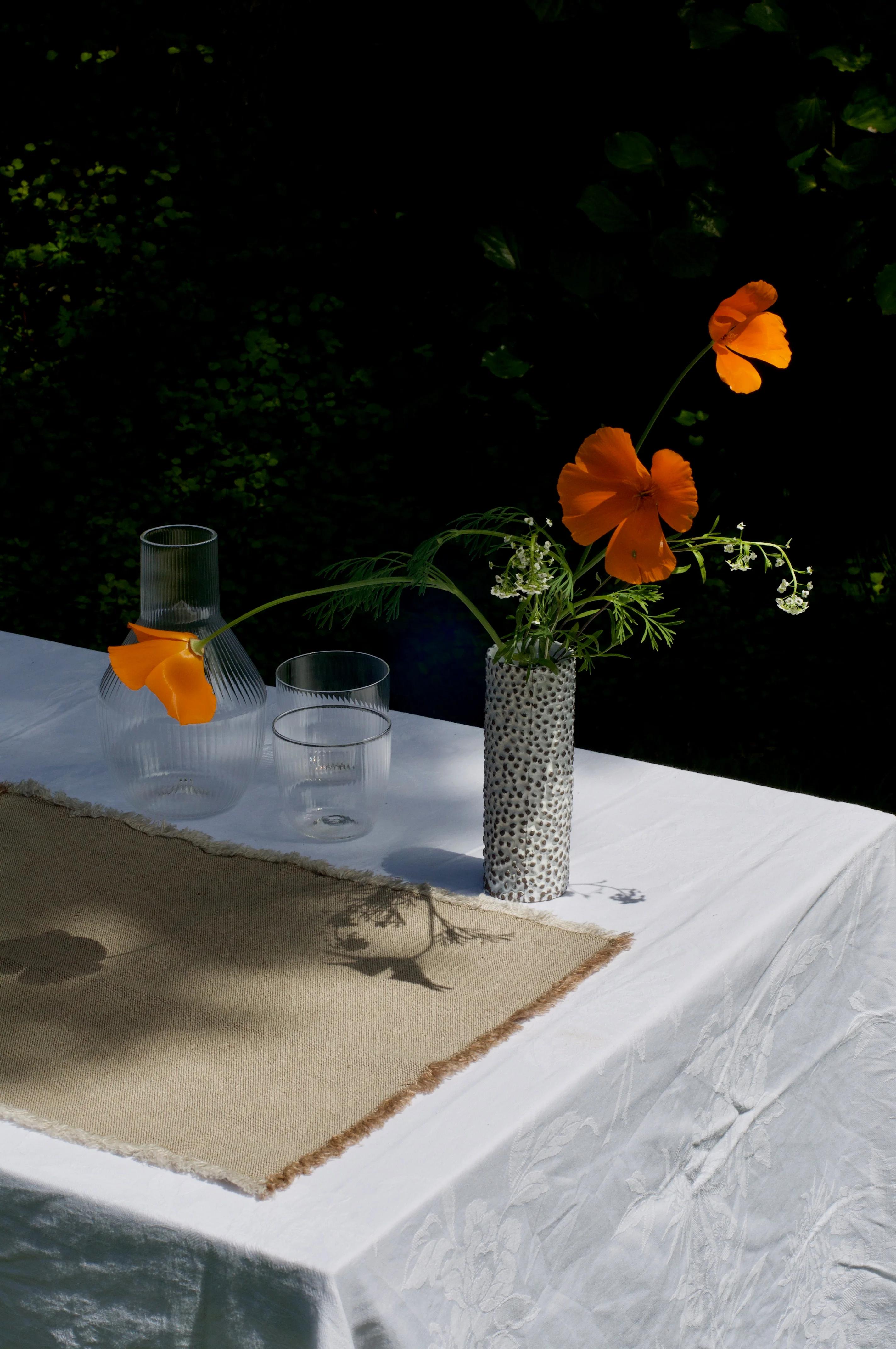 Outdoor table with clear vases and speckled vase holding orange flowers. Beige fabric adds contemporary touch. Green foliage backdrop.