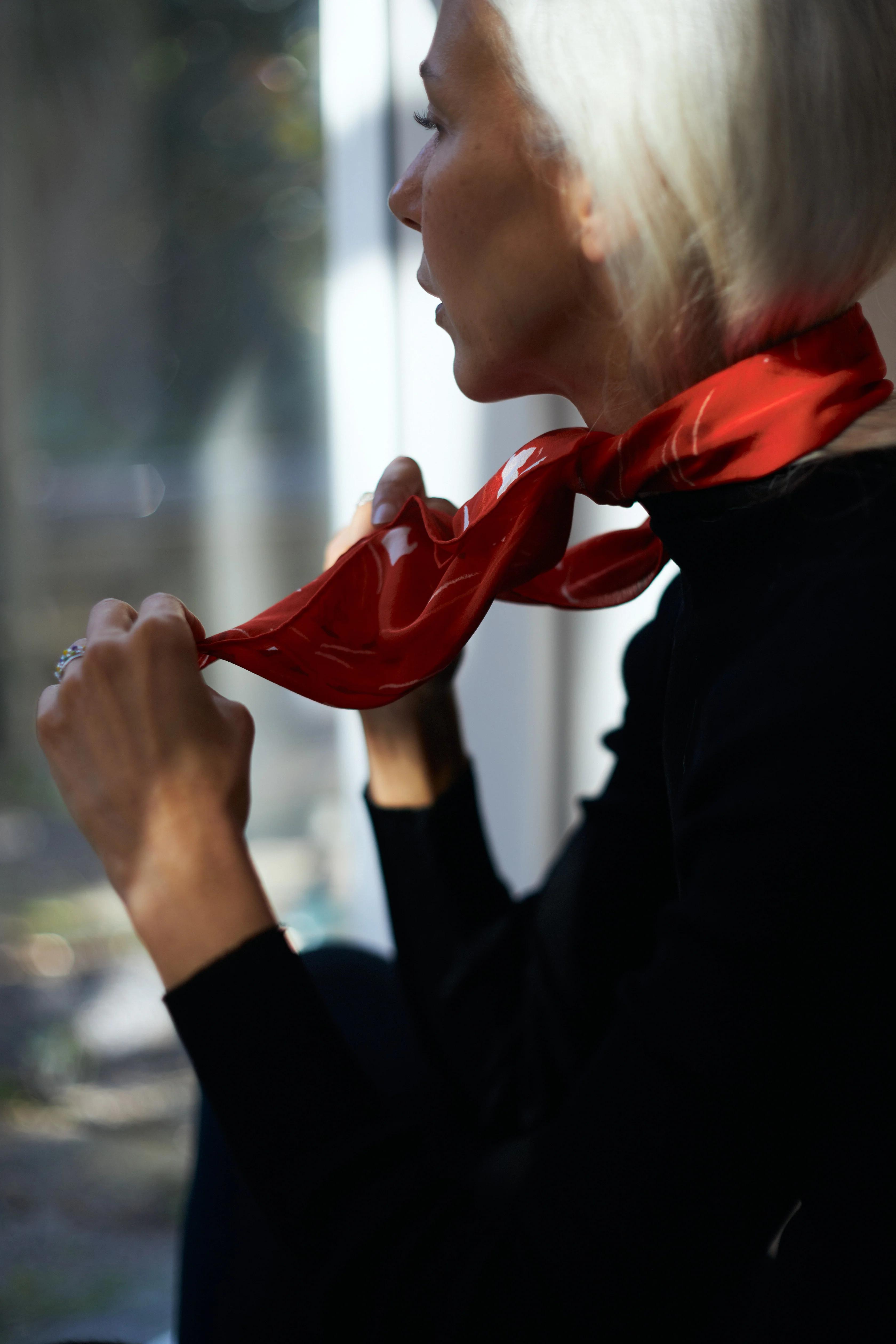 A person with white hair is tying a red scarf around their neck. Dressed in a black top, they are partially silhouetted against a softly lit background, creating a serene and contemplative atmosphere.