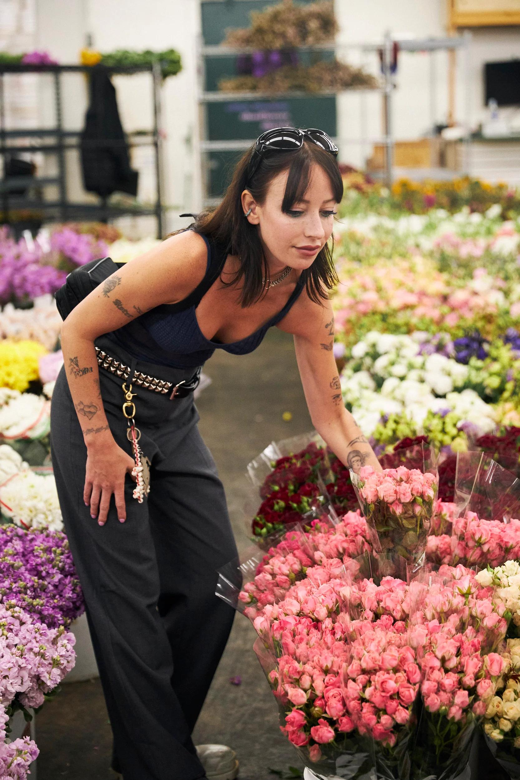 Person with black hair reaches for pink roses in flower-filled room. Navy tank top, dark pants, arm tattoos.