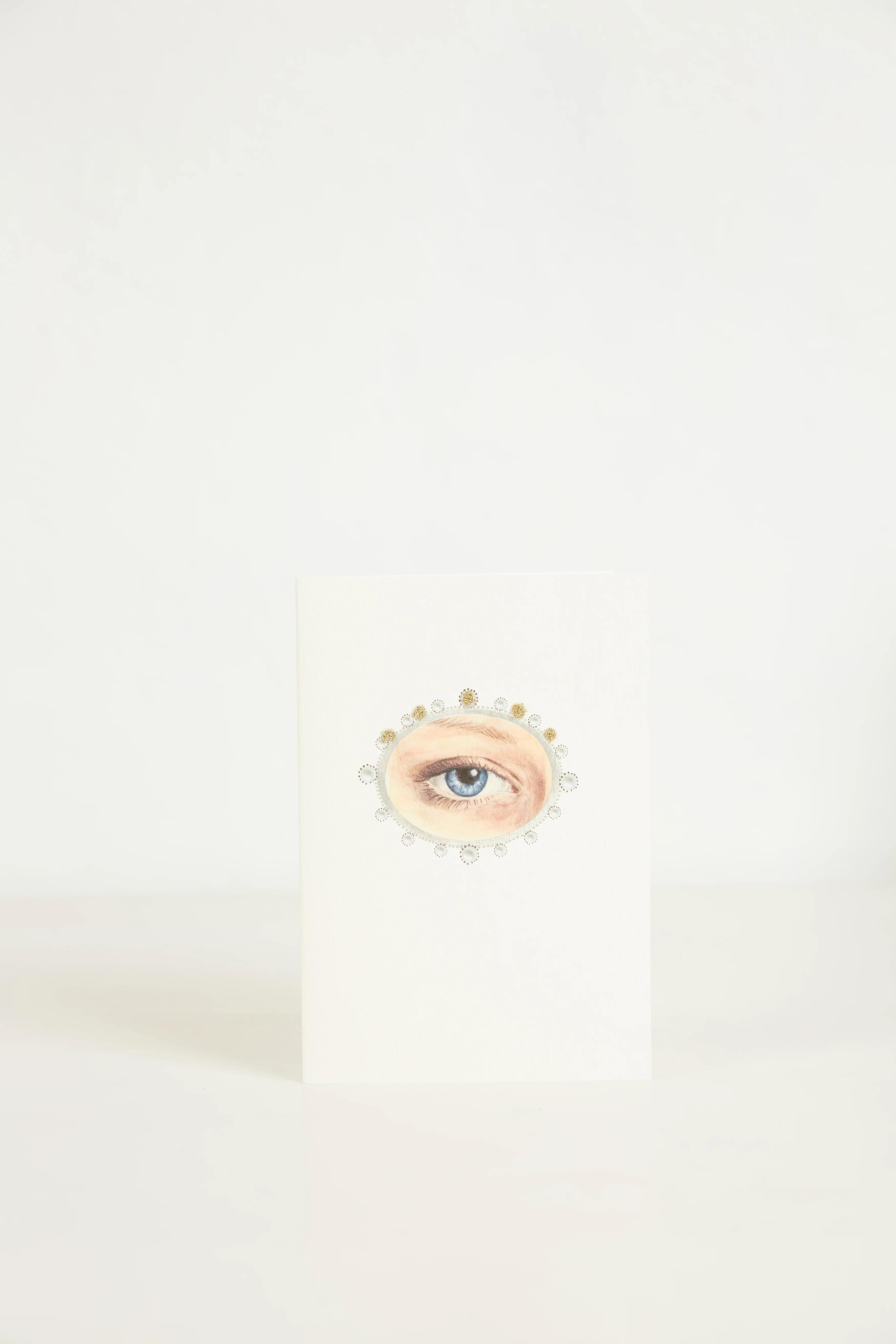 A greeting card with a drawing of an eye and surround encircled by small decorative elements. The card stands against a plan white background.