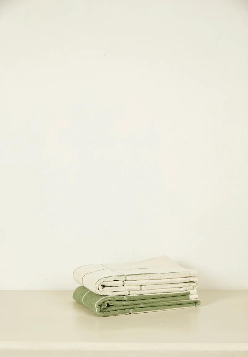 Two folded towels, one green and one beige, from the Antipodean brand Baina, placed on a white surface. The minimalist background emphasizes the clean and luxurious presentation.