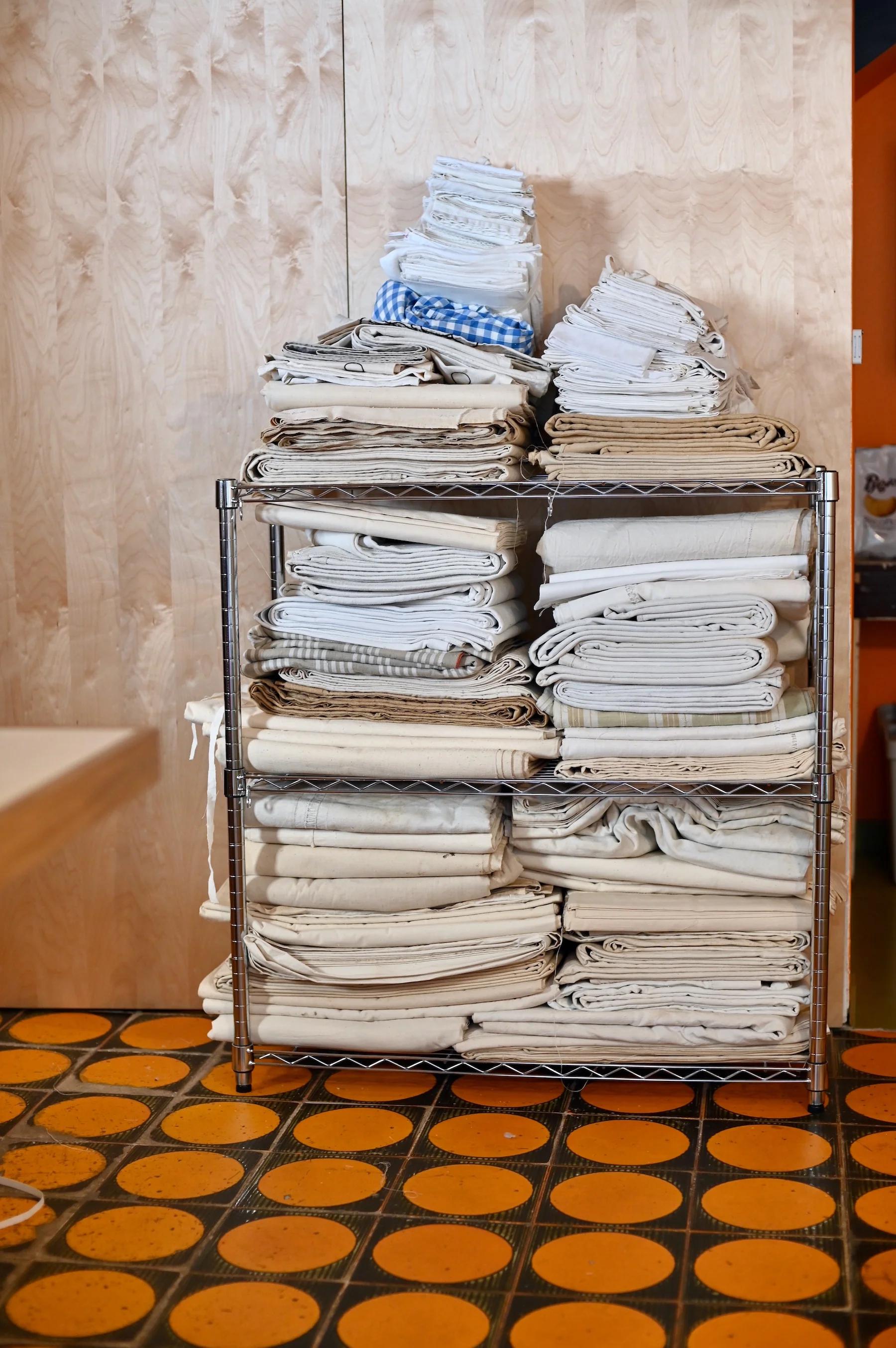 Metal rack with stacked neutral-coloured linens. Wooden wall and patterned floor in background.