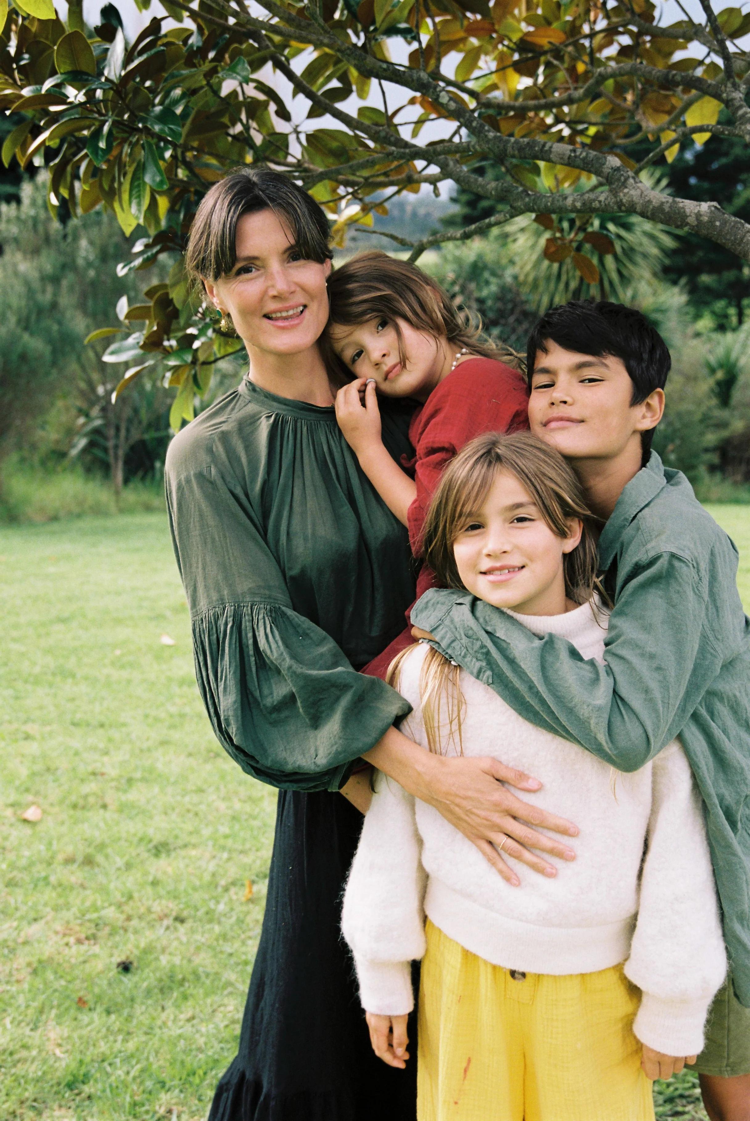 A woman in a dark green dress hugs three children under a tree. One child in red rests on her shoulder, another in yellow pants stands in front, and a third hugs her from the side.