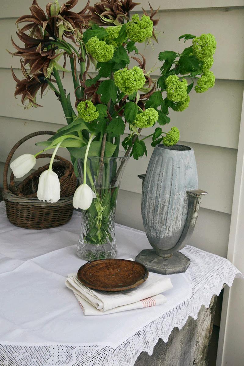 Holiday table with lace cloth, white tulips, and decorative items. Wooden trays on folded linens. White wooden wall background adds charm.