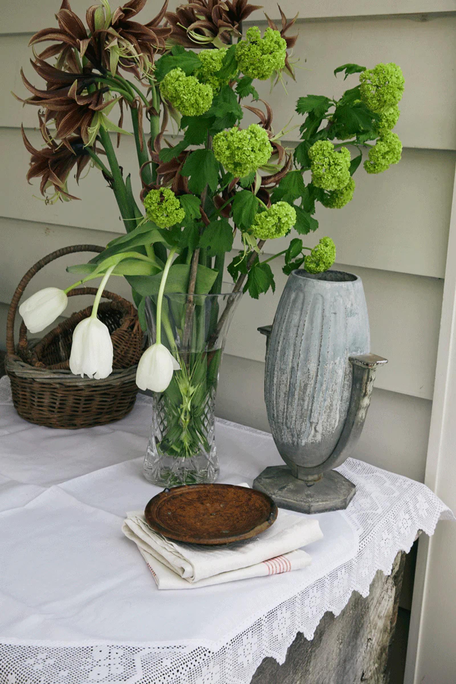 Holiday table with lace cloth, white tulips, and decorative items. Wooden trays on folded linens. White wooden wall background adds charm.