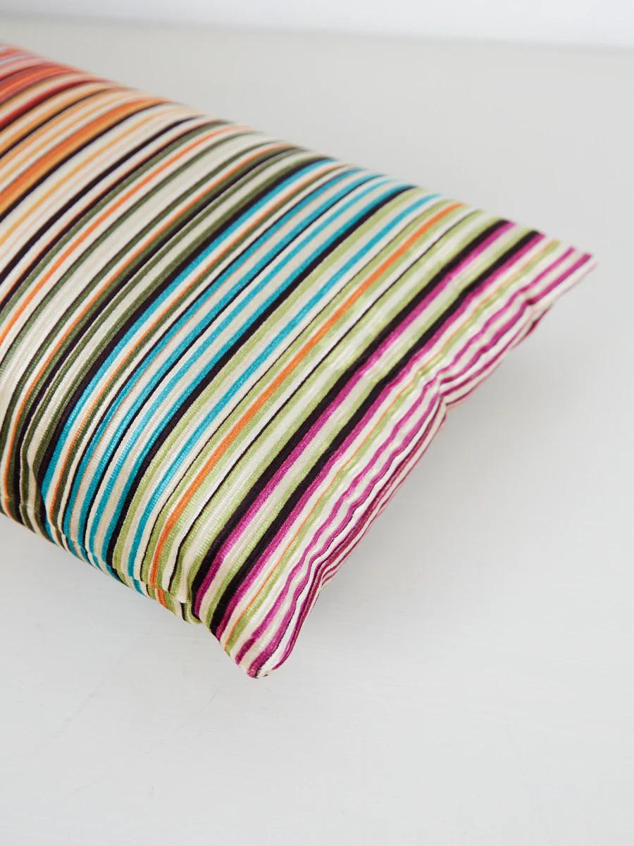 Rectangular striped pillow. Orange, brown, green, blue, pink, white. Light-coloured background.