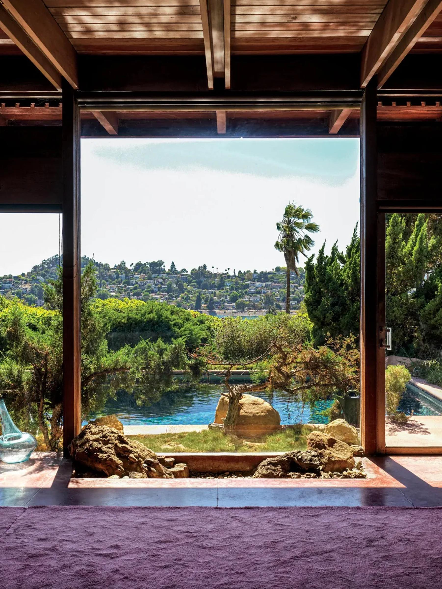 A serene landscape view from inside a room shows a lush garden with large rocks and pool in the foreground. A palm tree stands by the water with distant hillsides with houses in the background.