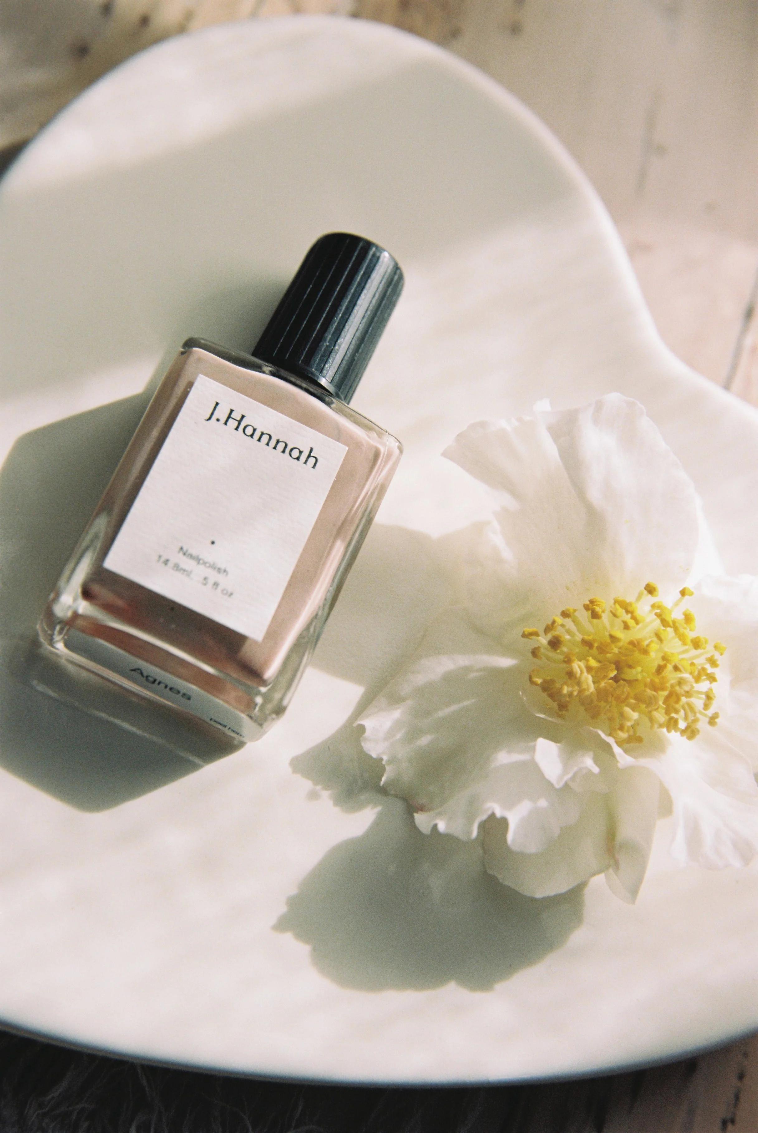 A glass bottle of J. Hannah nail polish lies on a heart-shaped ceramic dish next to a large white flower with a yellow centre. Soft, natural light creates an elegant, serene ambiance.