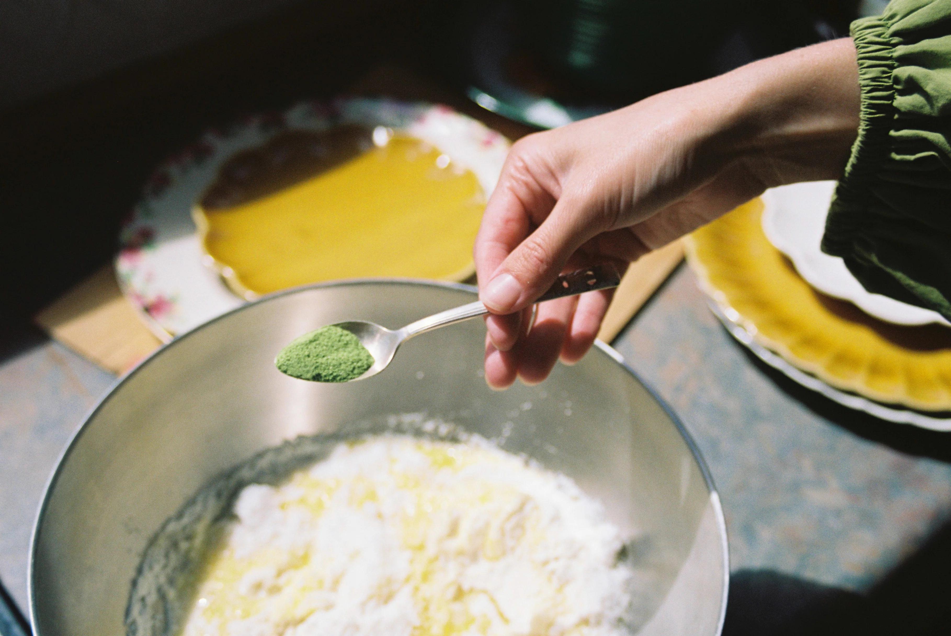 Hand with spoon of green powder over mixing bowl. Floral plate with yellow substance nearby. Person wearing green sleeve.