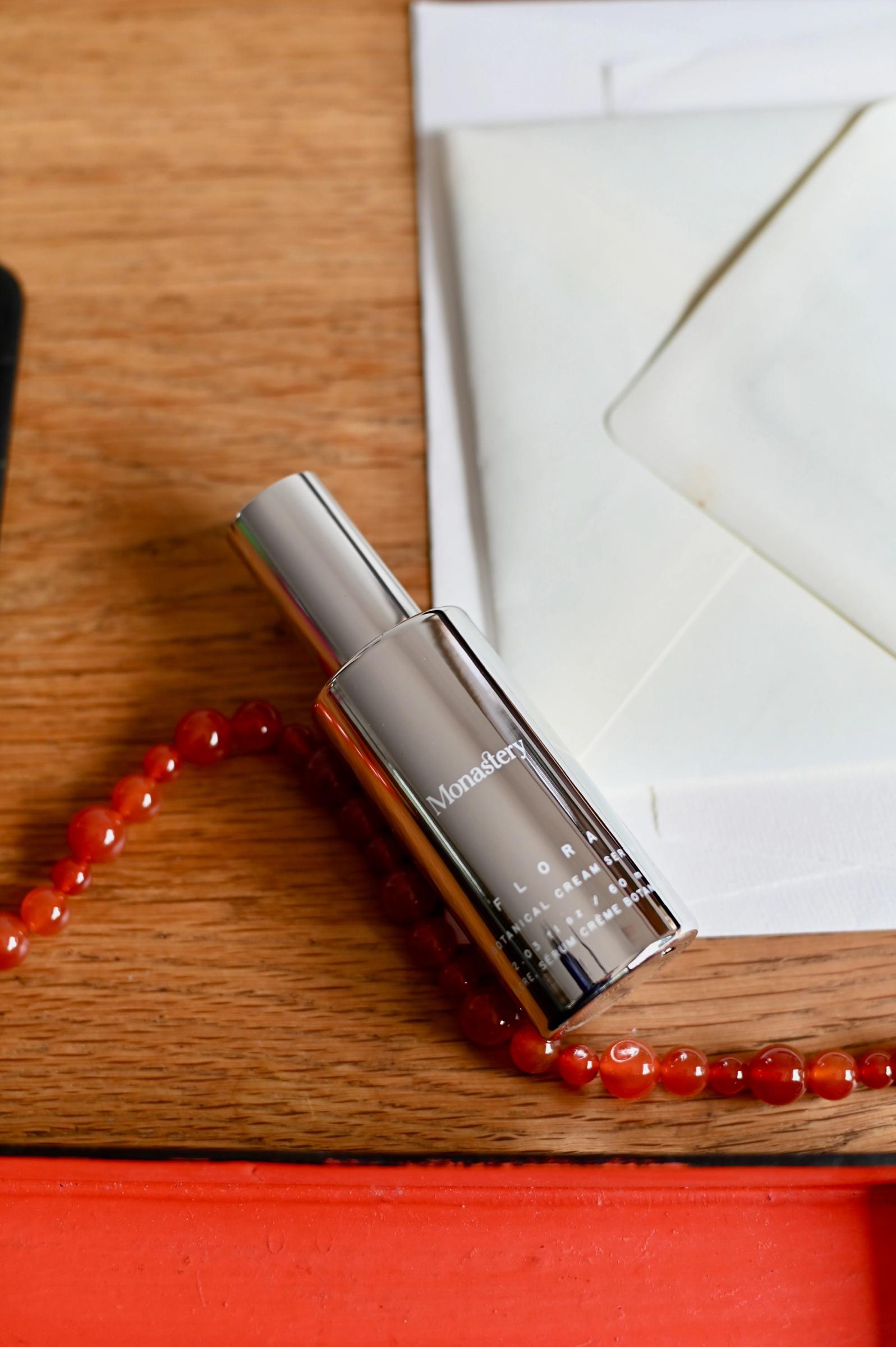 Silver bottle of Monastery Flora Botanical Cream Serum next to its brown box on light surface against neutral background.