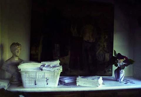 Dimly lit room with marble bust, laundry basket, and vase of red flowers. Dark painting on wall behind.