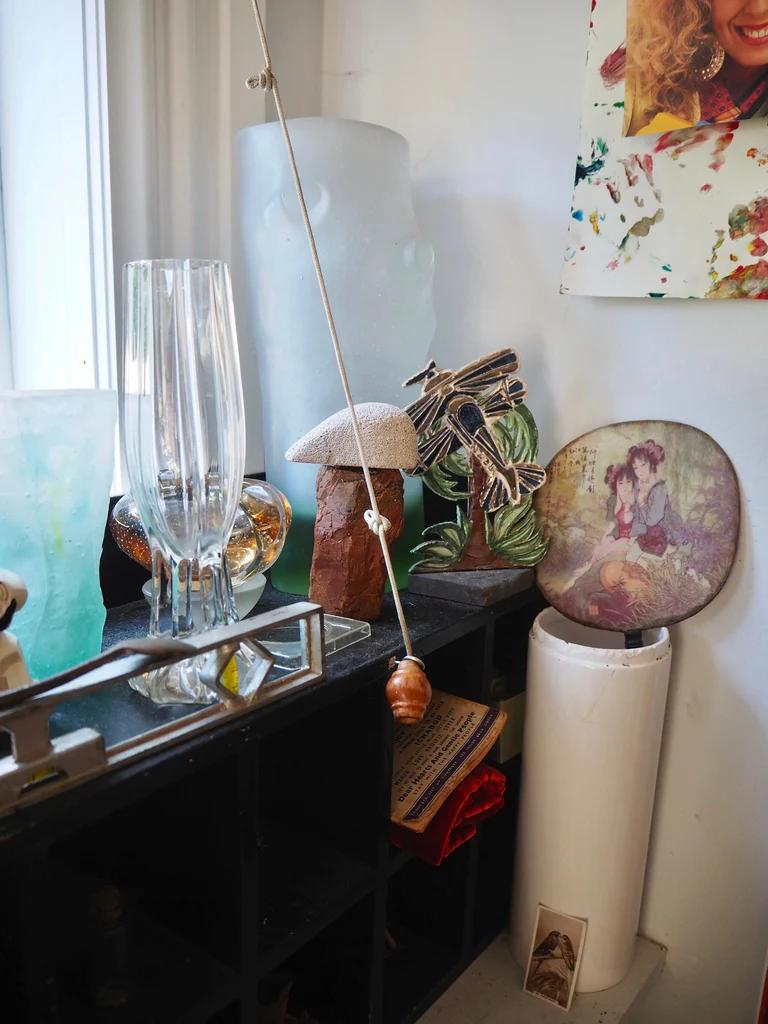 Eclectic shelf display with glass vases, ornaments, and photos. Translucent head sculpture and rolled papers. Hanging snail shell creates museum-like feel.