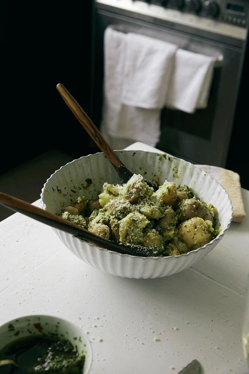 A white bowl of pasta topped with cheese sits on a white table. Wooden utensils and a dish with green sauce are nearby, reflecting seasonal Tessuti family recipes.