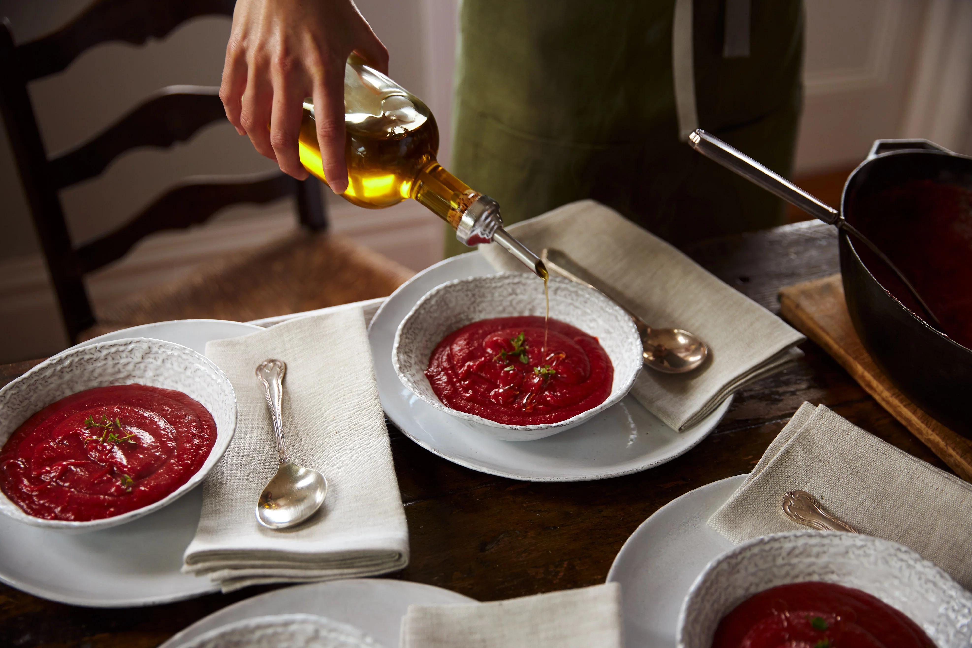 A person in a green apron pours olive oil into a bowl of beetroot soup garnished with herbs, surrounded by similar bowls and a ladle on a wooden table.