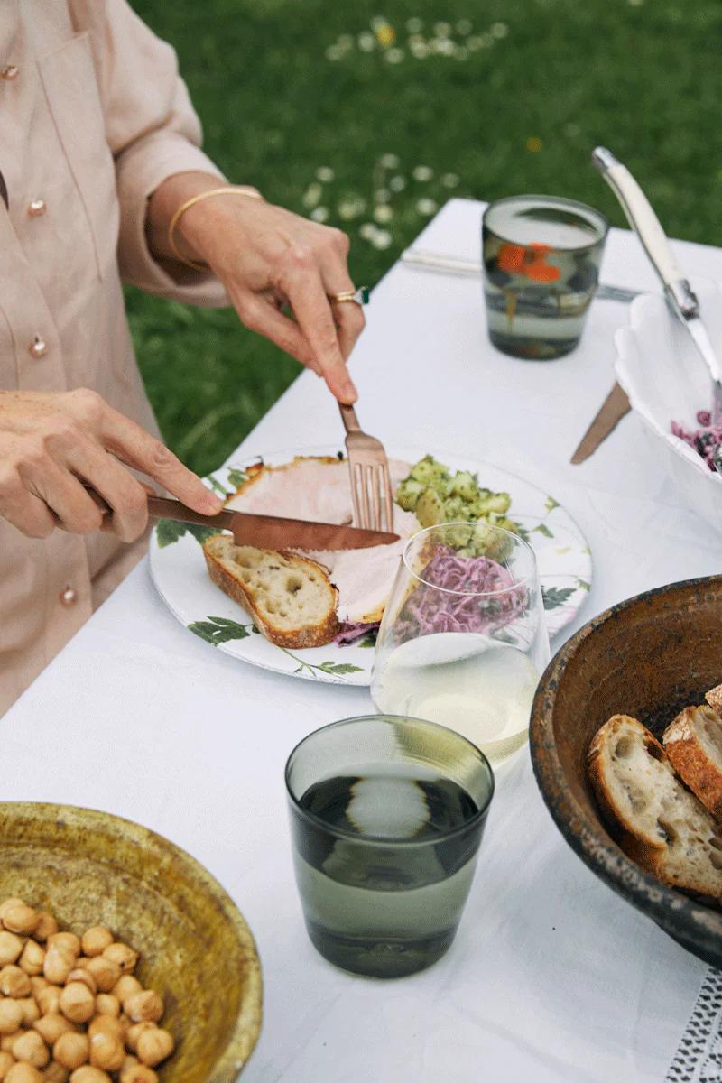 Person in beige shirt eating at flower-adorned table. Plate with meat and vegetables, glasses and dishes visible.