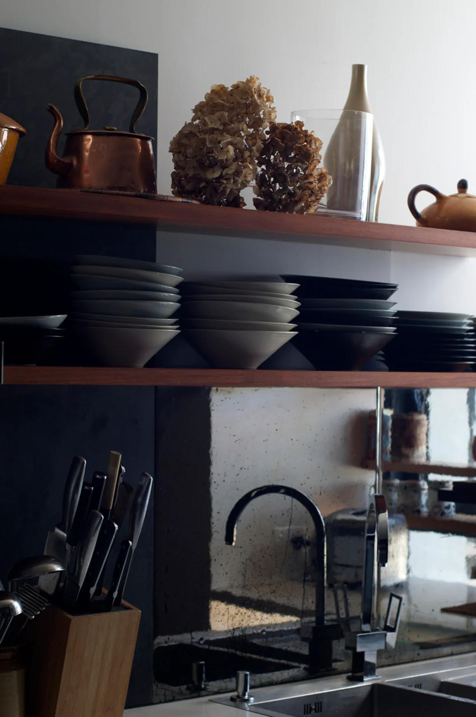 Kitchen with wooden shelves holding bowls, plates, and copper kettle. Counter features knife block and modern faucet. Walls have reflective, rustic backsplash.