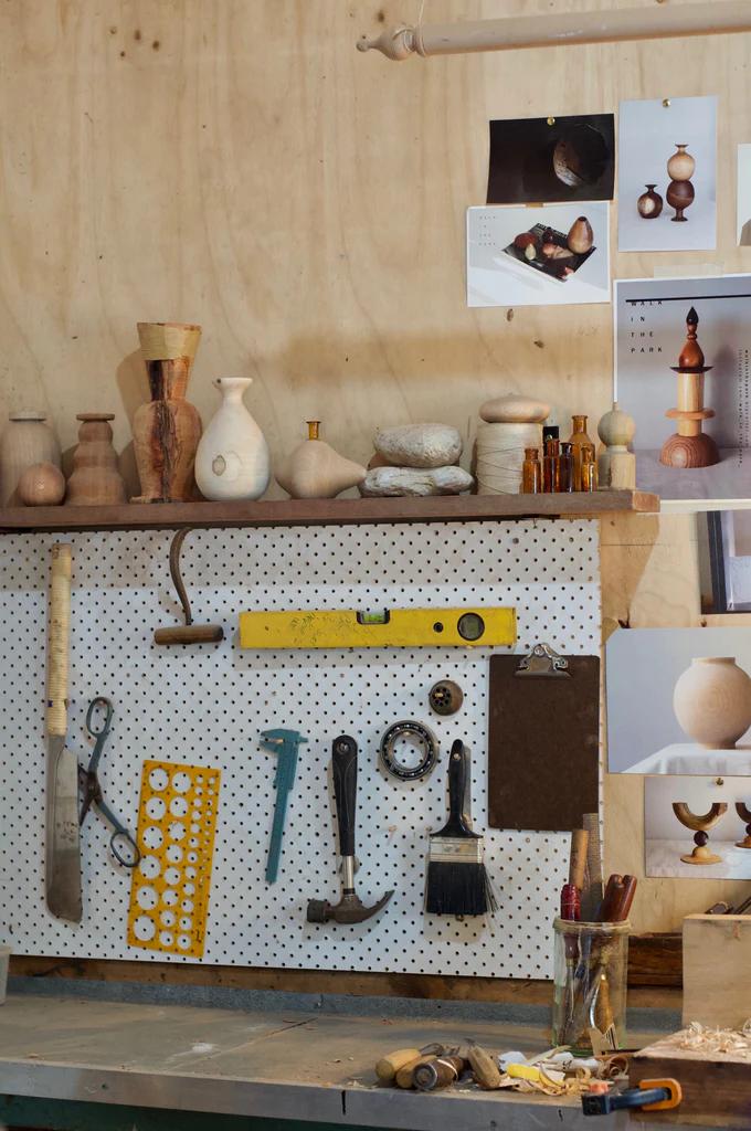 Woodworking bench with pegboard displaying tools. Shelves hold wooden vases, wall shows pinned photos and sketches of NZ wood products.