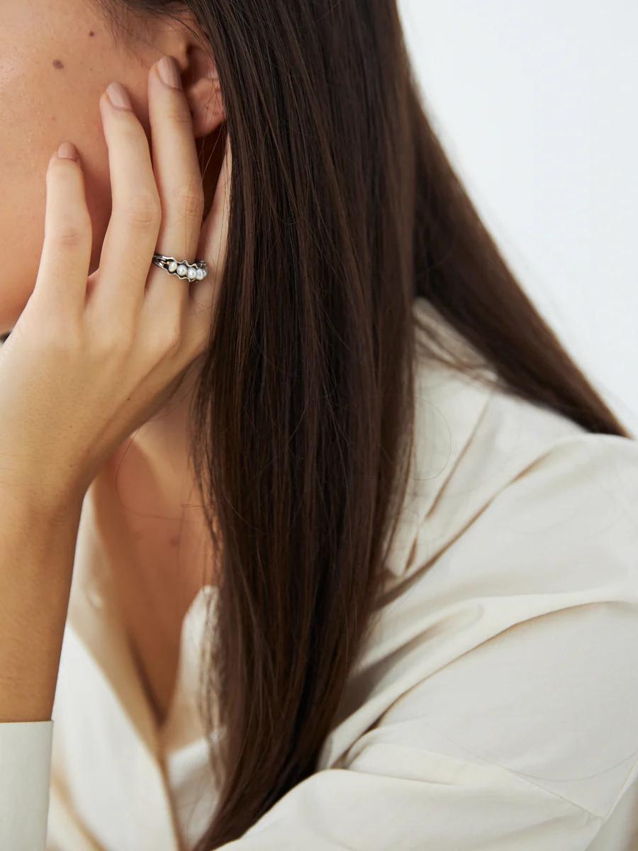 Woman with long brown hair rests hand on face, revealing multiple silver rings. Wears light-coloured, long-sleeved shirt.