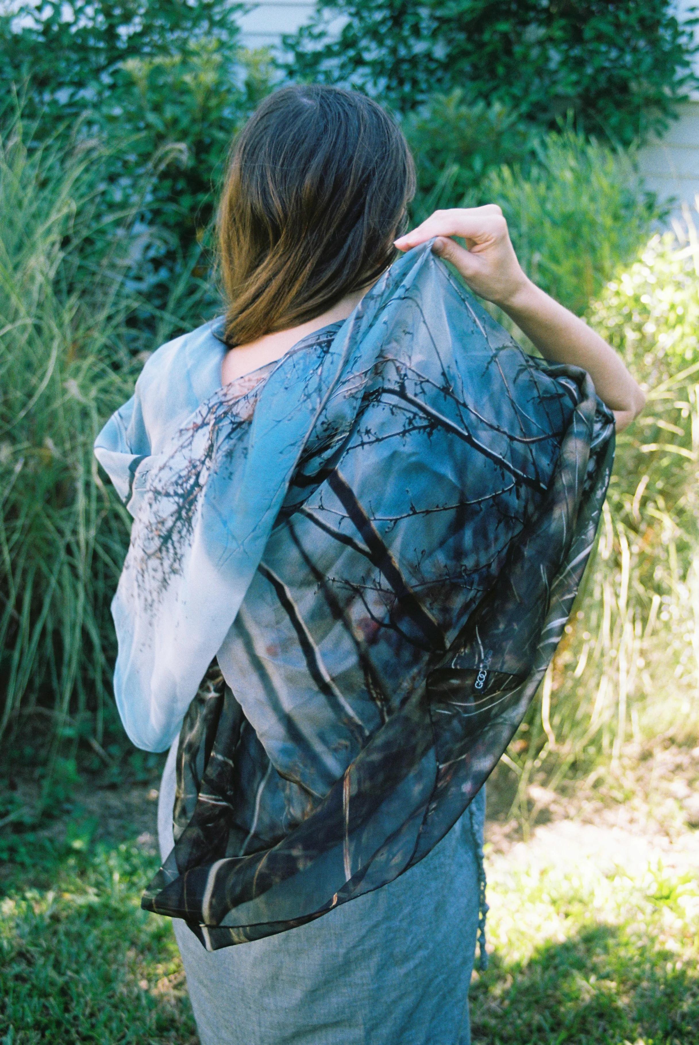 Long-haired person facing away, holding sheer shawl with tree branch design. Green plants and grass in background.