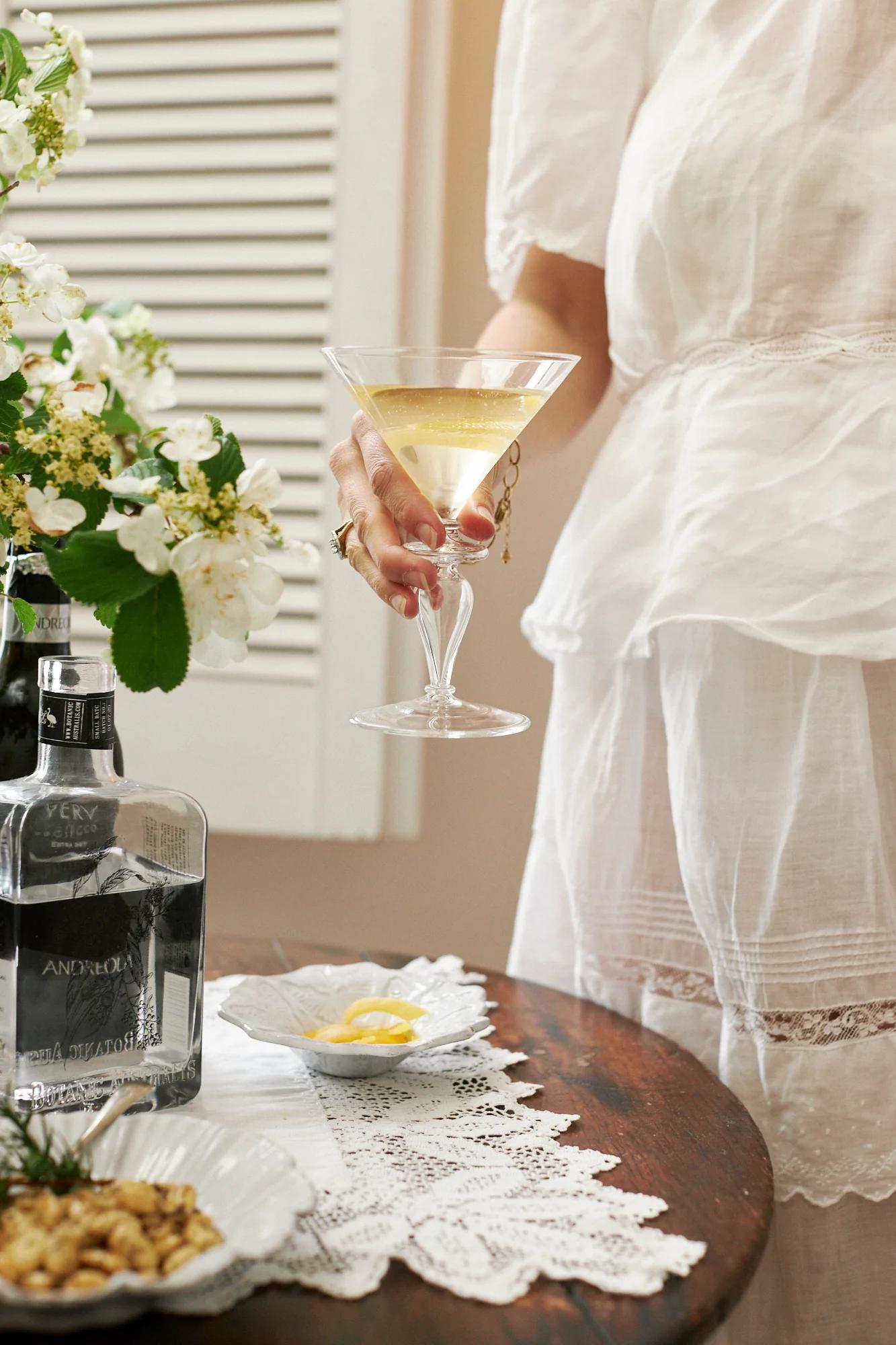 A person in a white dress holds a martini. The table has vodka, lemon twists, and snacks. A floral arrangement and lace runner complete the elegant setting.