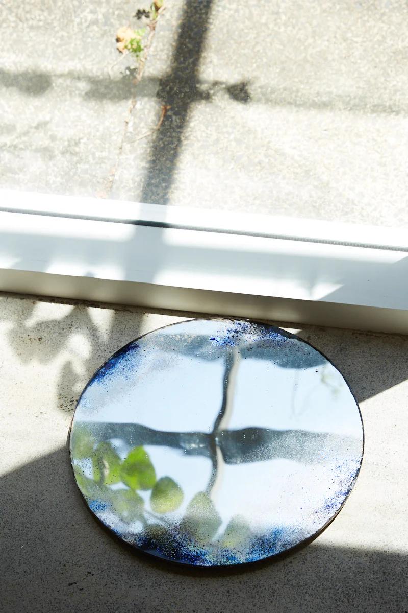 Round metallic mirror with blue border near window. Reflects light and window frame. Shadows on floor.