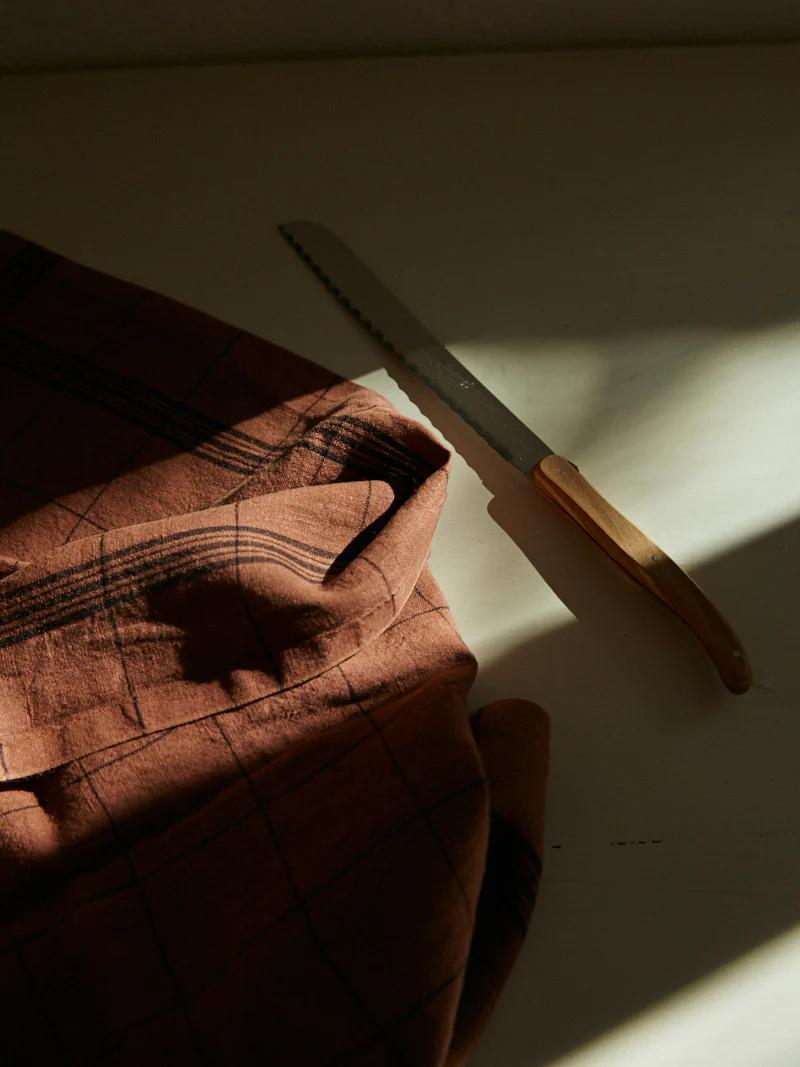 Serrated knife near plaid cloth partially covering bread loaf. Natural light casting soft shadows.