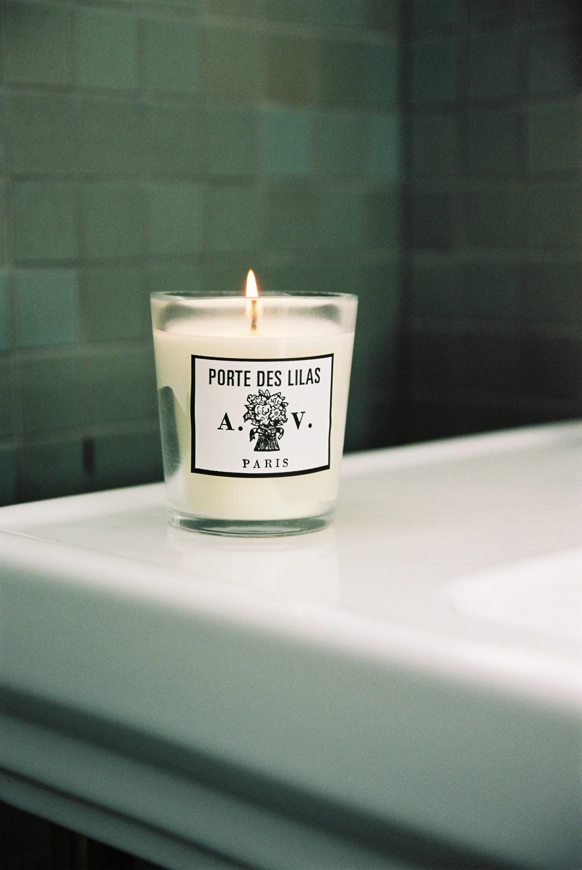 A lit candle in a glass container sits on a white bathroom sink. The label reads “Porte des Lilas, A.V. Paris” with an illustration of flowers. Light green tiles are in the background.