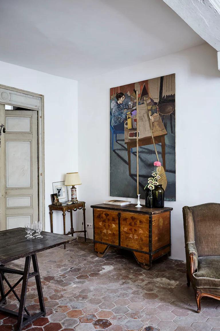 Cosy living room with hexagonal tiles and antique wooden dresser. Framed painting, side table with lamp, and upholstered chair create a charming Parisian ambiance.