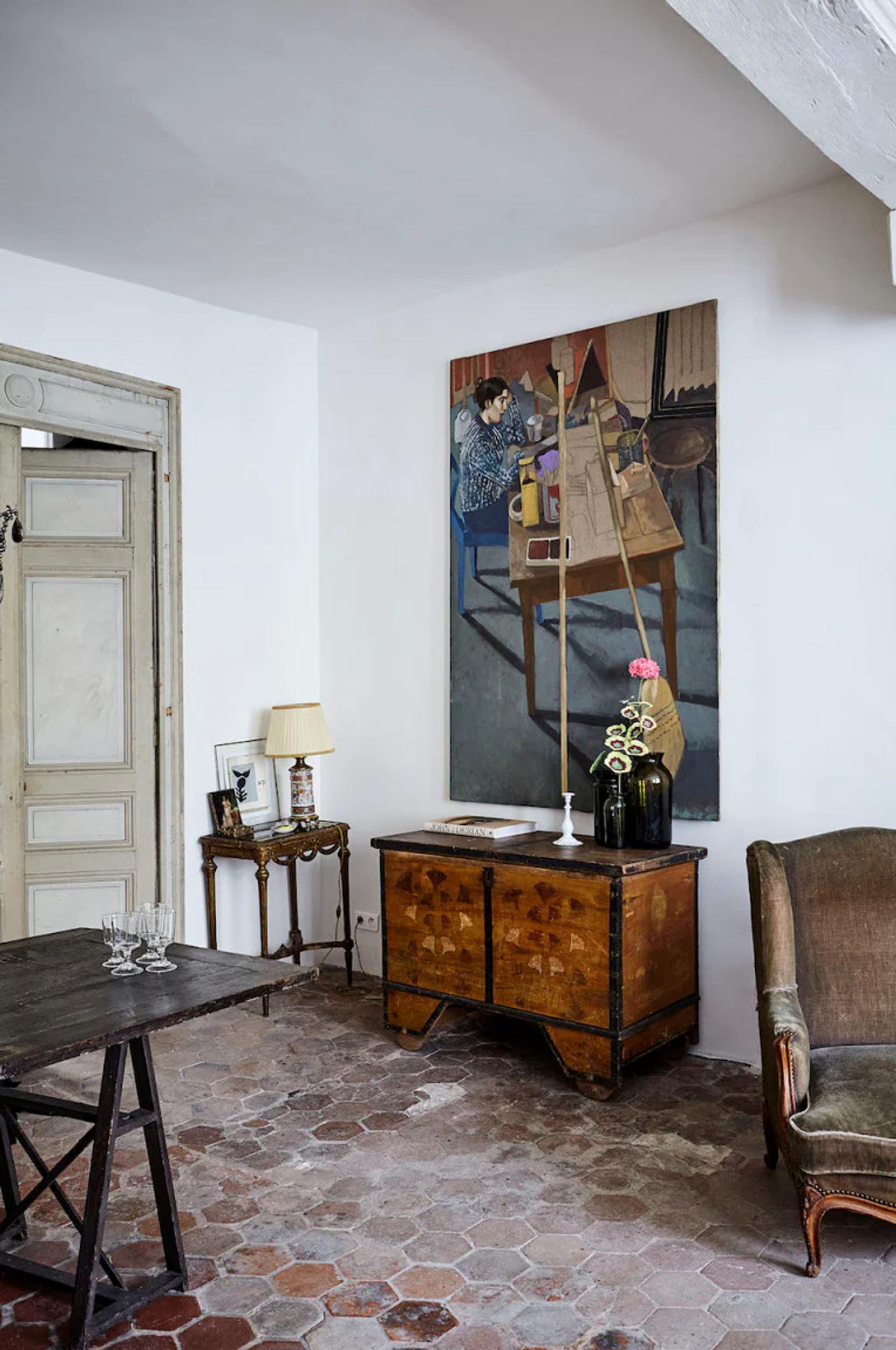 Cosy living room with hexagonal tiles and antique wooden dresser. Framed painting, side table with lamp, and upholstered chair create a charming Parisian ambiance.