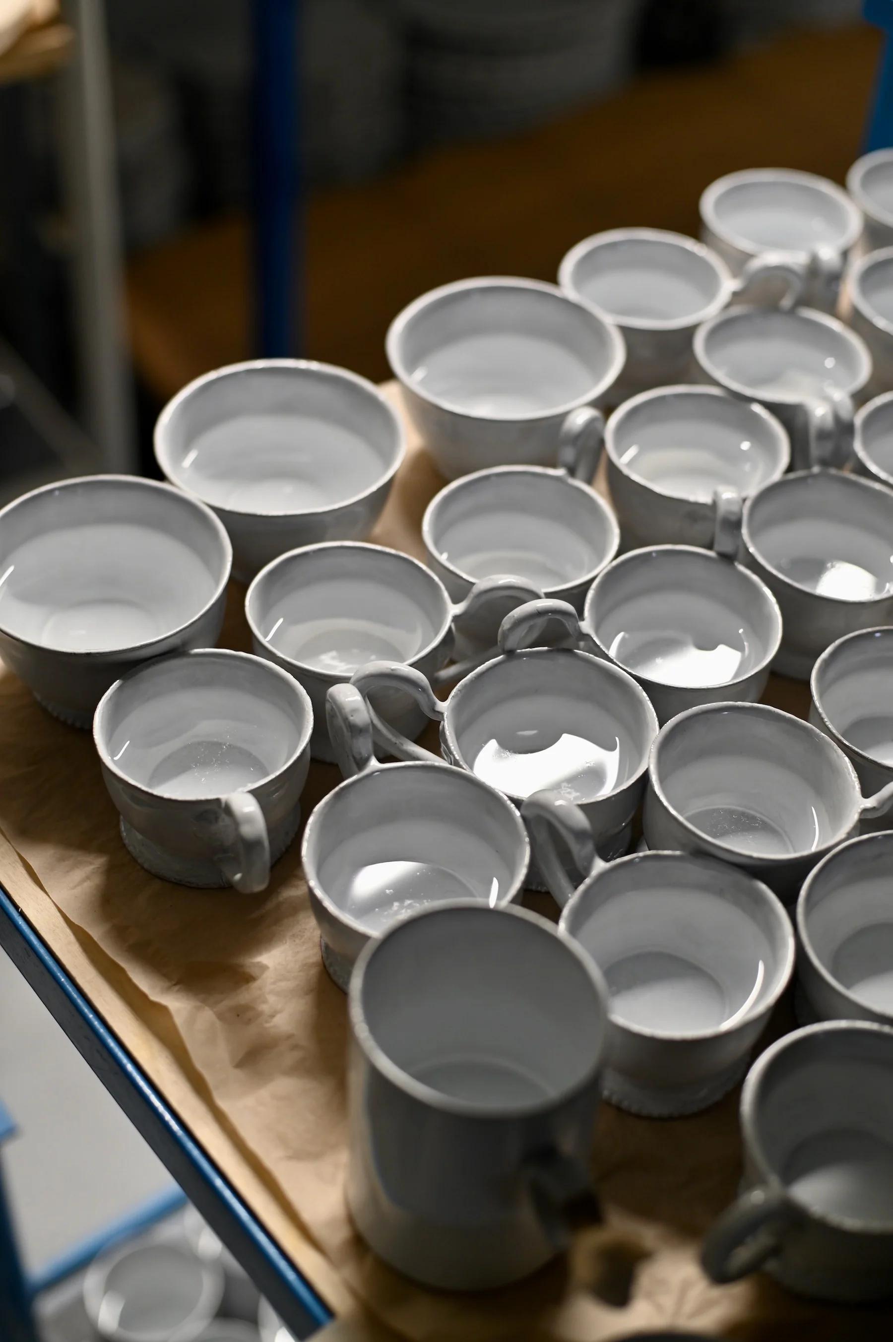 Collection of white ceramic cups with handles on brown paper-covered surface. Cups in various stages of completion, some filled with water.