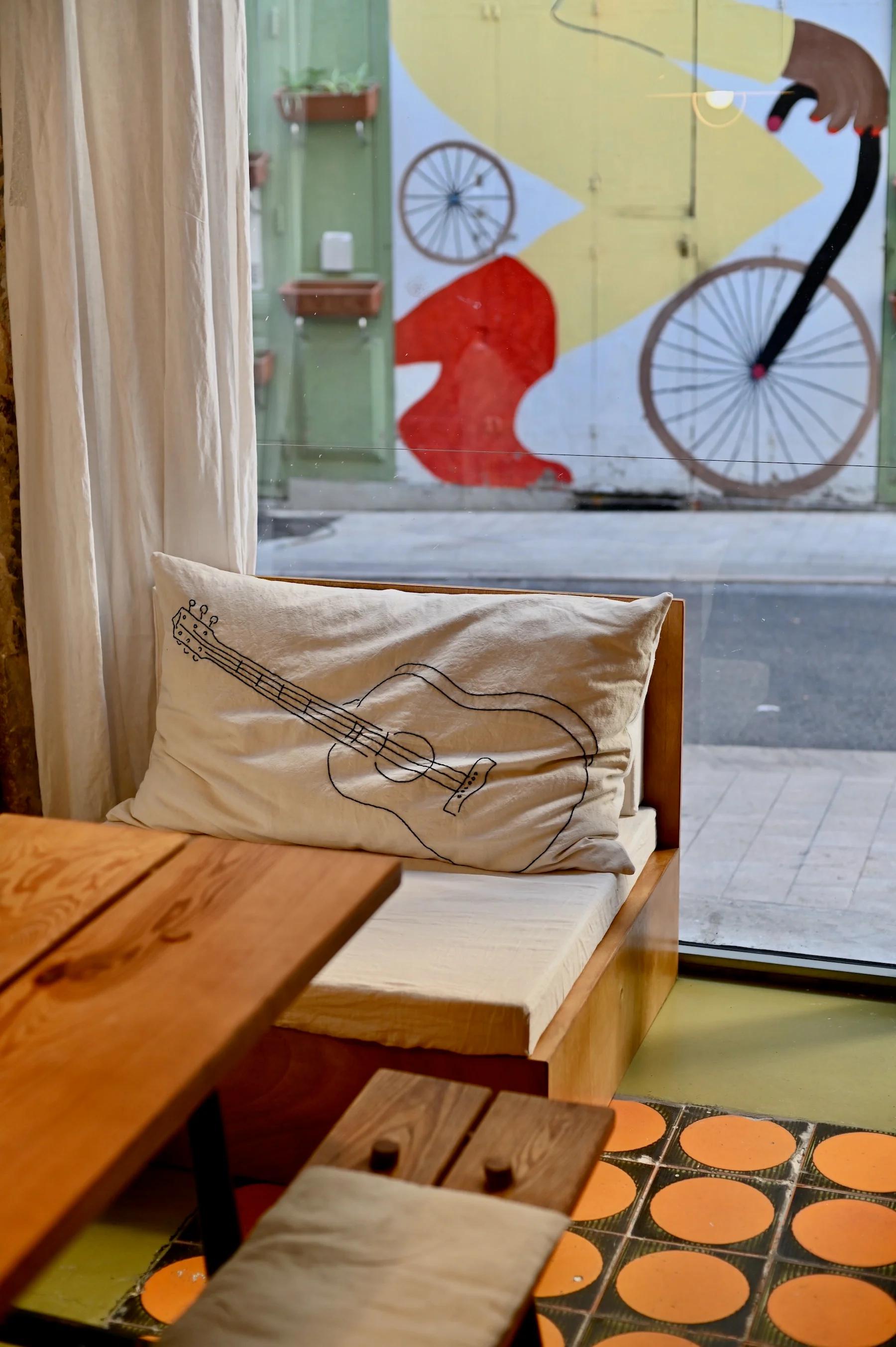 Wooden table and benches with cushions near window. Colourful mural visible outside. Pillow with guitar sketch on bench.