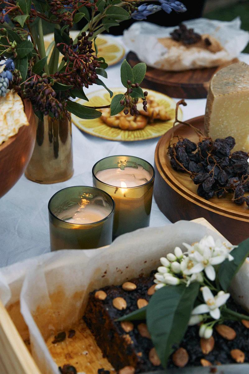 Table with assorted foods, candles, flowers, and Christmas cake. Vase with floral arrangement in background.