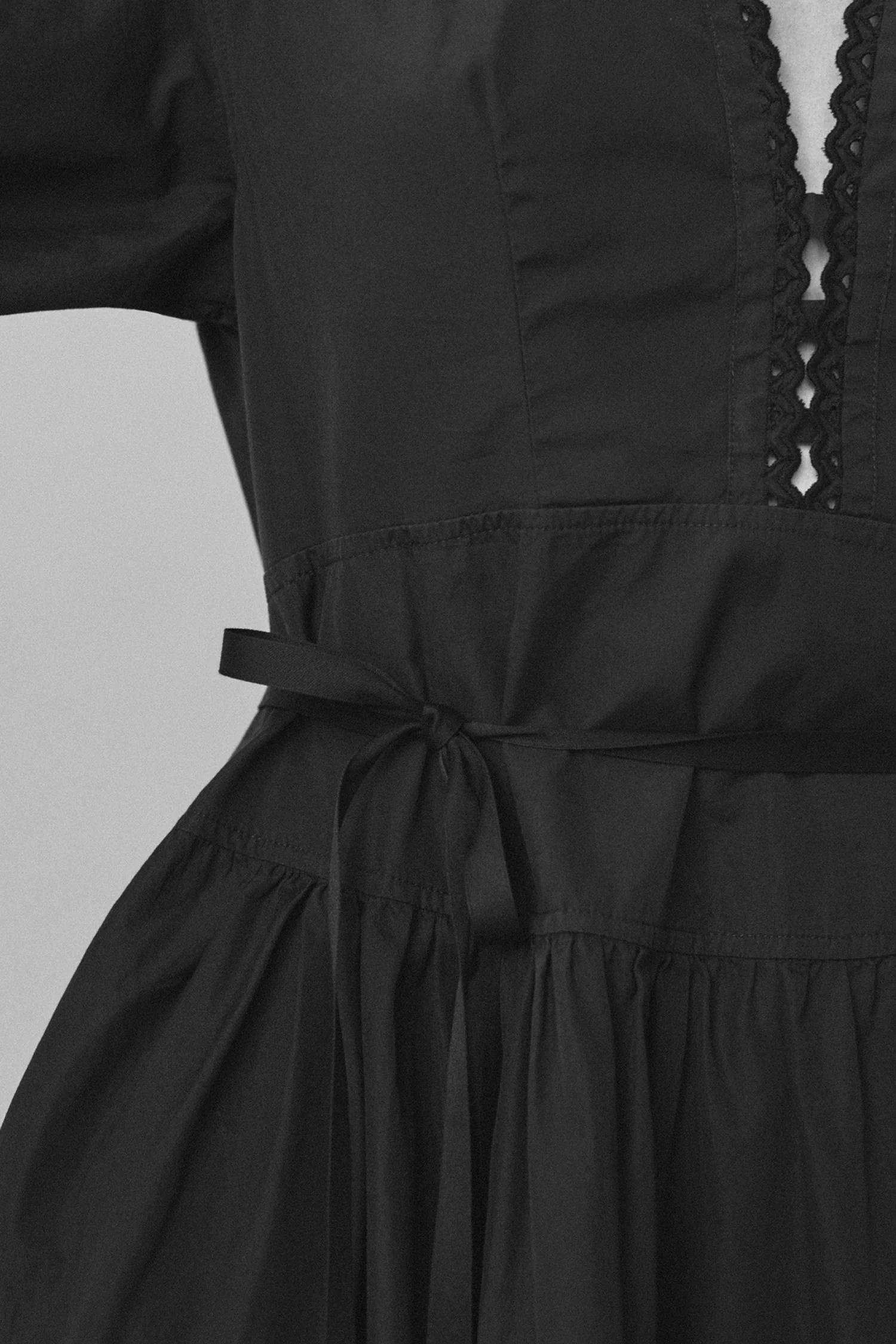 Close-up of black dress with detailed neckline and tied waist. Long sleeves, fitted bodice, gathered skirt.