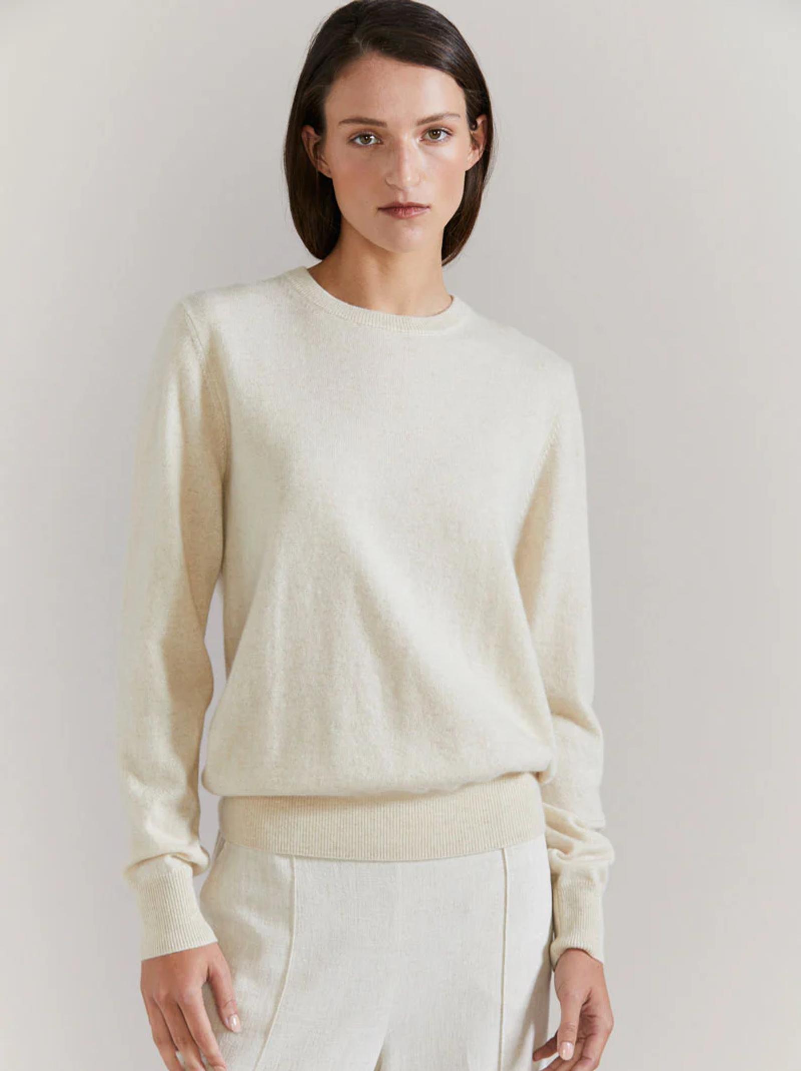 Person in light-coloured sweater and pants. Standing against plain background, looking at camera with neutral expression.