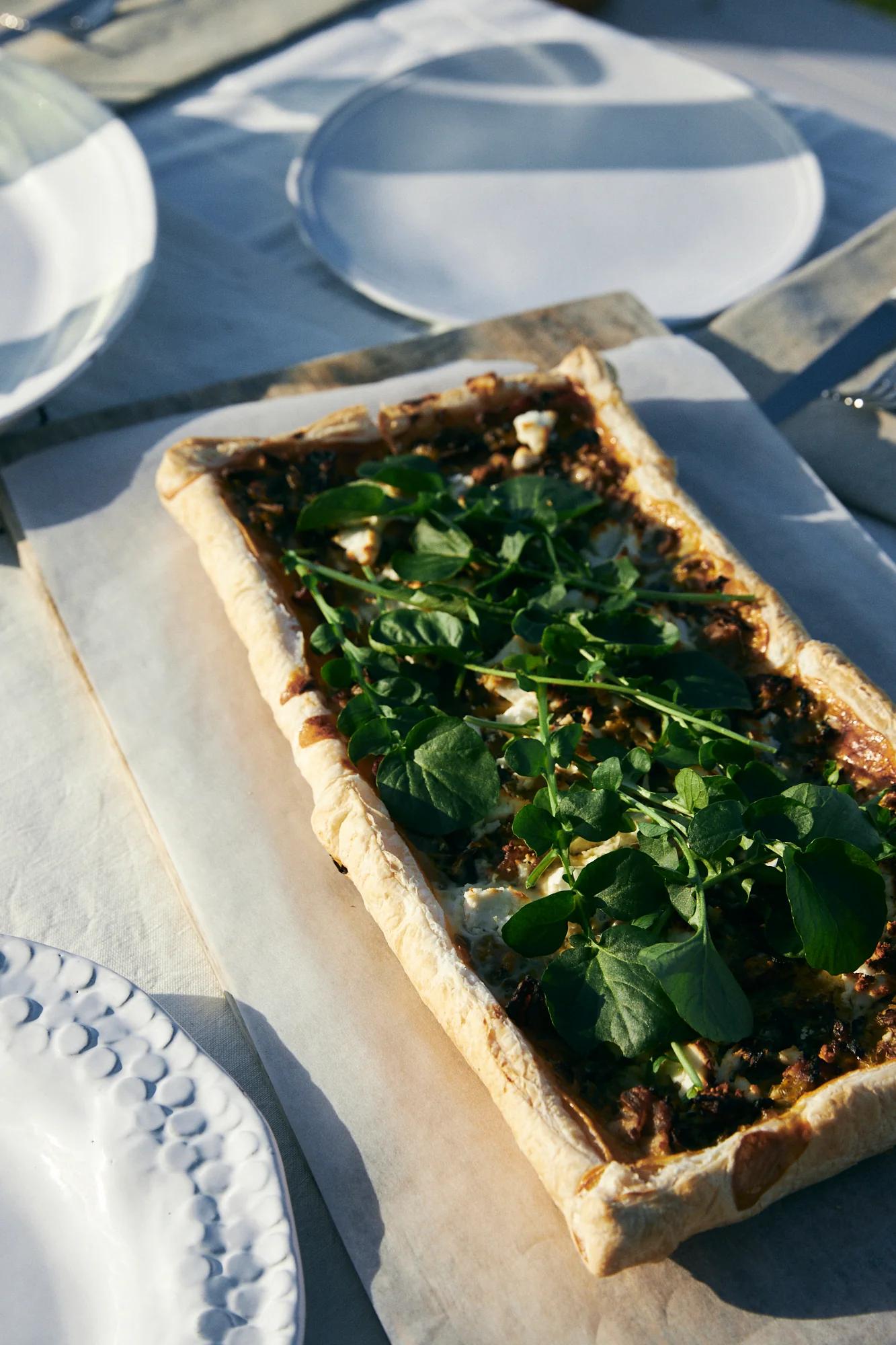 Rustic tart with greens and goat's cheese on parchment. Light tablecloth, white plates and cutlery nearby.