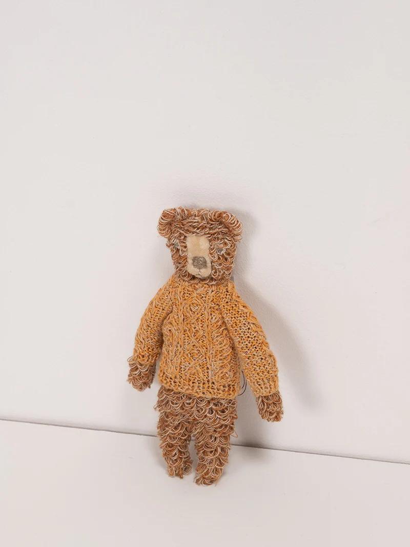 A curly-haired teddy bear wearing a hand-crochet orange sweater. The bear’s fur is light and dark brown, creating a textured look against a white background.