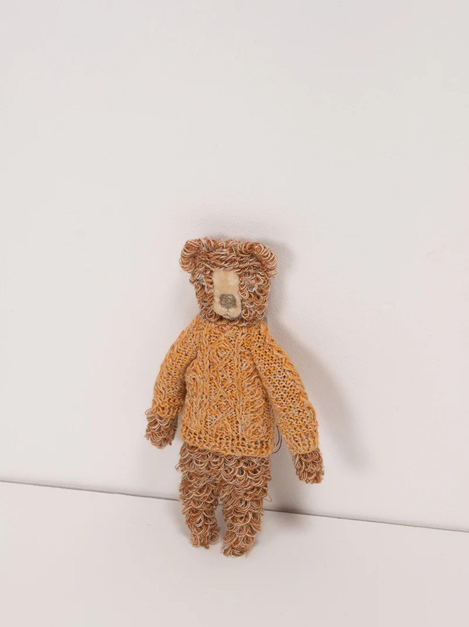 A curly-haired teddy bear wearing a hand-crochet orange sweater. The bear’s fur is light and dark brown, creating a textured look against a white background.