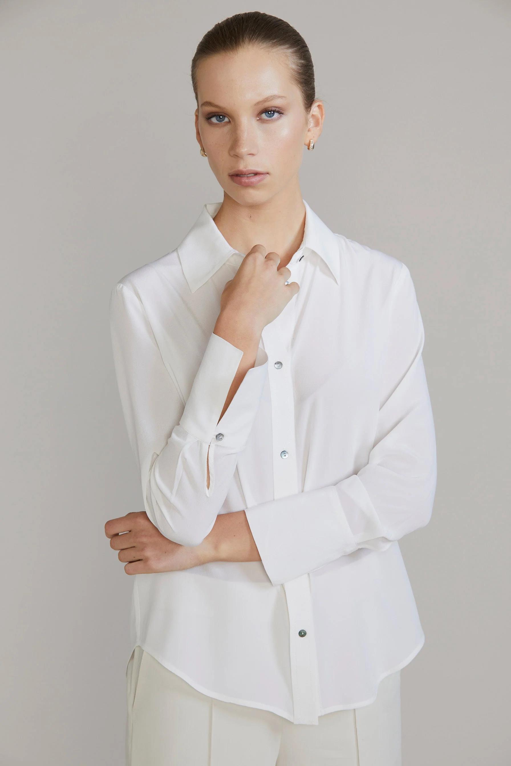 Woman in white button-up shirt and cream trousers. One hand resting on opposite wrist. Plain background.