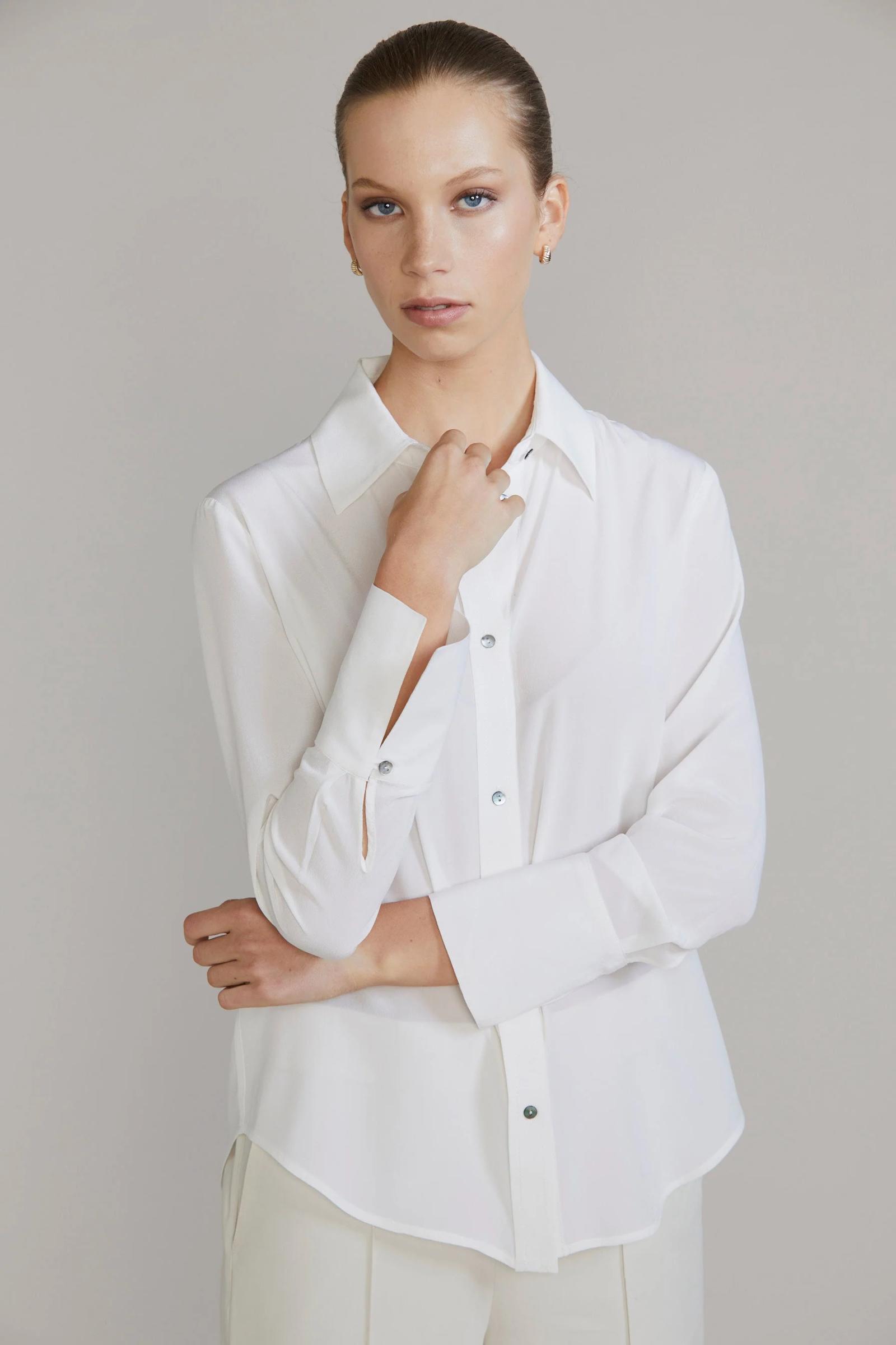 Woman in white button-up shirt and cream trousers. One hand resting on opposite wrist. Plain background.